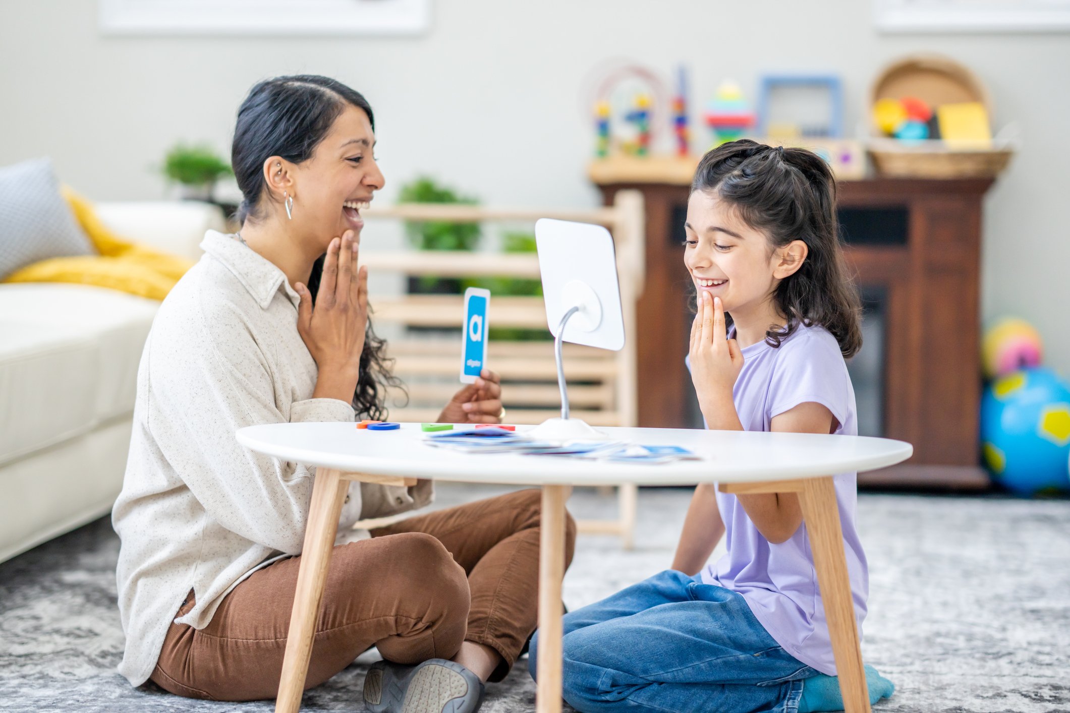 A female speech therapist works with a young girl as they practice their sounds and letter enunciation. The Therapist is dressed casually and is holding up alphabetical cue cards as they work together.
