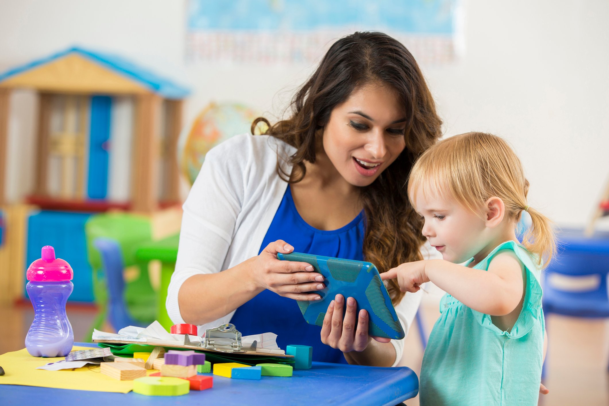 Helpful Hispanic female teacher shows a cute blonde Caucasian toddler something on a digital tablet. They are sitting at a table in a daycare learning center working on the tablet.